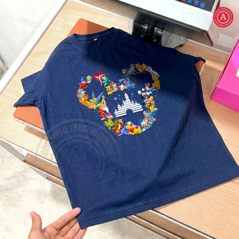 Peut inclure: T-shirt bleu marine avec un motif t&ecirc;te de Mickey Mouse compos&eacute; de divers personnages Disney et d'une silhouette de ch&acirc;teau. Le t-shirt a des manches courtes et est fait d'une mati&egrave;re douce. Le texte "AURORA FINDS SHOP" est &eacute;galement visible.