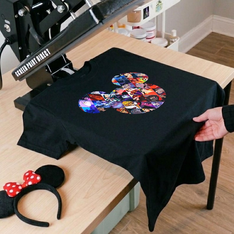 Puede incluir: Camiseta negra con un dise&ntilde;o de cabeza de Mickey Mouse lleno de im&aacute;genes de personajes de dibujos animados coloridos. Tambi&eacute;n se ve una diadema con lazo de lunares rojos y blancos y una m&aacute;quina de prensado en caliente.
