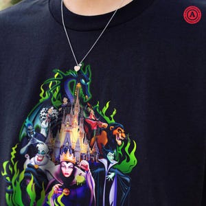 Puede incluir: Camiseta negra con un gr&aacute;fico colorido de villanos de Disney, incluyendo Mal&eacute;fica y &Uacute;rsula, con un castillo de fondo y llamas verdes. Se ve un collar plateado con un colgante de coraz&oacute;n.