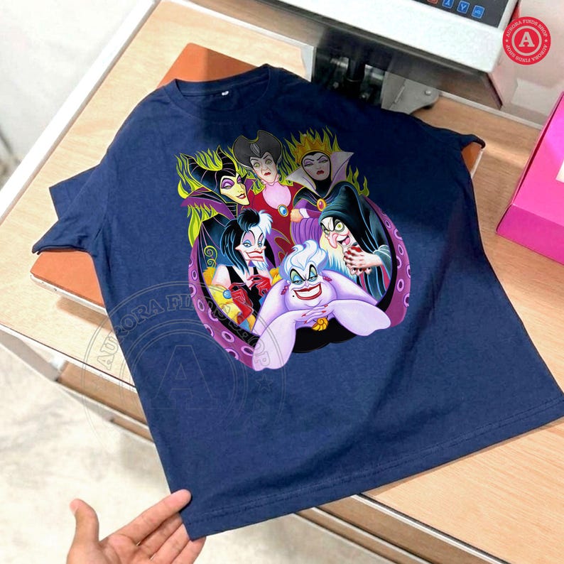May include: Navy blue t-shirt featuring a colorful graphic of Disney villains, including Ursula, Maleficent, and the Evil Queen. The design is set within a purple and pink circular frame. The shirt is made of a soft, dark blue fabric.