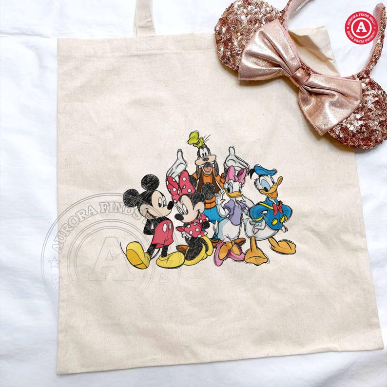 May include: A cream canvas tote bag with a colourful graphic of Mickey Mouse, Minnie Mouse, Goofy, Daisy Duck, and Donald Duck. A pair of sequined rose gold mouse ears with a bow are next to the bag.