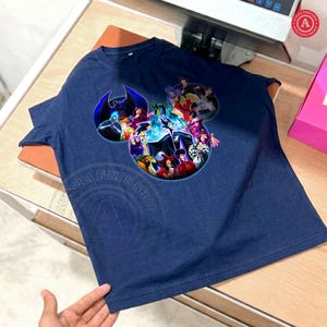 K&ouml;nnte beinhalten: Marineblaues T-Shirt mit einem farbenfrohen Aufdruck von Disney-B&ouml;sewichten, die in Form von Mickey Mouse-Ohren angeordnet sind. Die Grafik zeigt Charaktere wie Ursula, Maleficent und die b&ouml;se K&ouml;nigin. Das Shirt ist aus weichem Material.
