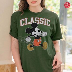 May include: Green t-shirt featuring a classic Mickey Mouse graphic. The word "CLASSIC" is printed above the character in white, distressed lettering. Mickey Mouse is depicted in his iconic pose, wearing orange shorts and yellow shoes.
