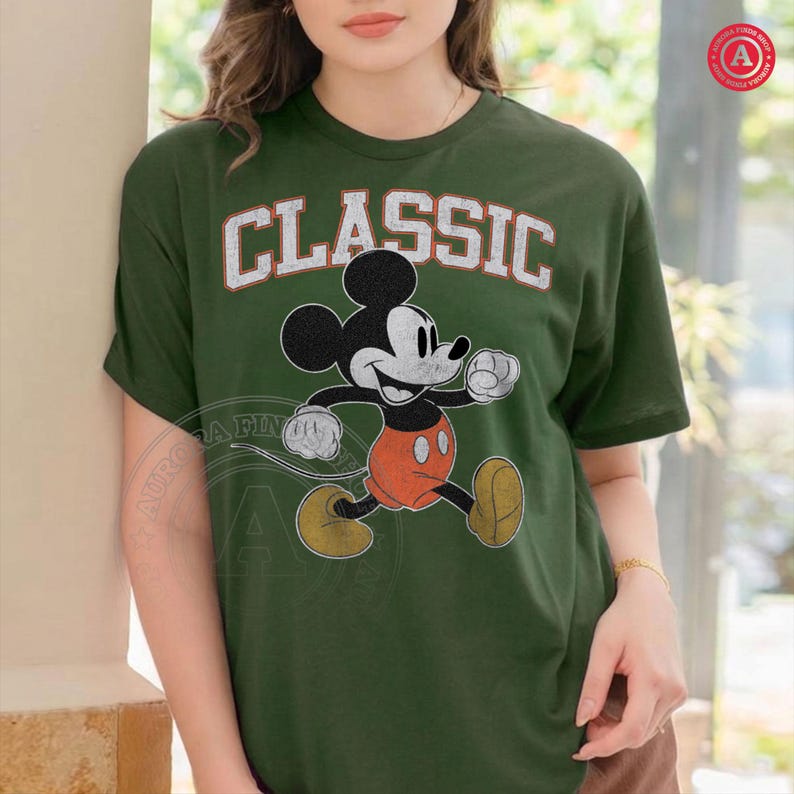 May include: Green t-shirt featuring a classic Mickey Mouse graphic. The word "CLASSIC" is printed above the character in white, distressed lettering. Mickey Mouse is depicted in his iconic pose, wearing orange shorts and yellow shoes.