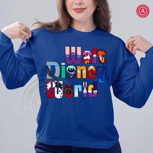 Puede incluir: Una sudadera de manga larga azul real con las palabras "Walt Disney World" en un dise&ntilde;o colorido y en capas. Las letras incorporan varias im&aacute;genes de personajes y parques de Disney, creando un gr&aacute;fico vibrante y llamativo.