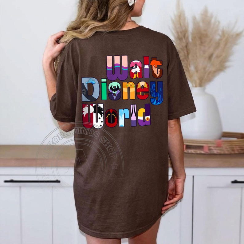 Puede incluir: Una camiseta marr&oacute;n con las palabras "Walt Disney World" en letras coloridas y estilizadas. Las letras est&aacute;n dise&ntilde;adas con varios temas de personajes de Disney. La camiseta tiene un corte casual y de gran tama&ntilde;o.