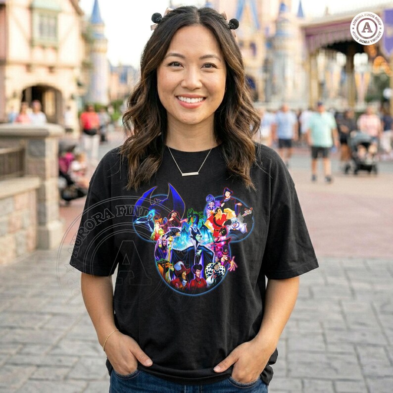 K&ouml;nnte beinhalten: Schwarzes T-Shirt mit einer farbenfrohen Grafik von Disney-B&ouml;sewichten in Form eines Mickey Mouse-Kopfes. Das Shirt hat einen Rundhalsausschnitt und kurze &Auml;rmel. Der Hintergrund zeigt einen Themenpark mit einem Schloss.