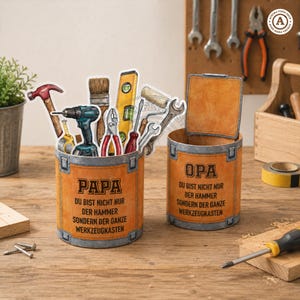 May include: Two orange-colored, cylindrical containers with the words "PAPA" and "OPA" printed on them. The "PAPA" container holds various tools, including a hammer, drill, and wrench. The "OPA" container is empty. The text on the containers reads "DU BIST NICHT NUR DER HAMMER SONDERN DER GANZE WERKZEUGKASTEN."