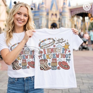 Op de afbeelding: Een wit t-shirt met korte mouwen en een kleurrijke grafische print met de tekst I KNEW IT I KNEW YOU en infinity and beyond. Het ontwerp bevat illustraties van een kralenarmband, cowboylaarzen, alfabetblokken en een hoed.