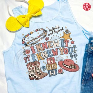 Op de afbeelding: Een lichtblauw hemdje met een grafische print met de tekst "I KNEW IT I KNEW YOU" en "infinity and beyond". Het ontwerp bevat een kralenketting, een ster, cowboylaarzen, alfabetblokken met "TSS 5" en een roze hoed met een koeienprint band.