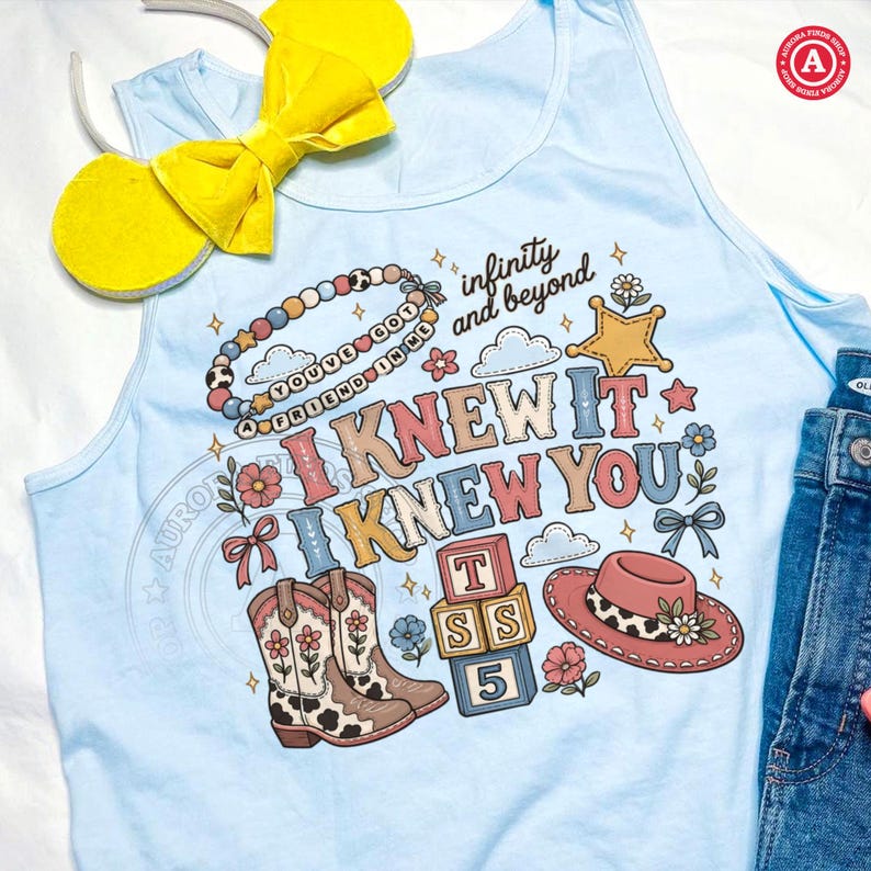 Op de afbeelding: Een lichtblauw hemdje met een grafische print met de tekst "I KNEW IT I KNEW YOU" en "infinity and beyond". Het ontwerp bevat een kralenketting, een ster, cowboylaarzen, alfabetblokken met "TSS 5" en een roze hoed met een koeienprint band.