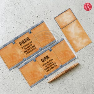 May include: A set of orange and gray paper cards with text in German, including "PAPA" and "OPA". The cards have a toolbox design. A wooden pen lies on the surface. The cards are on a light gray surface.