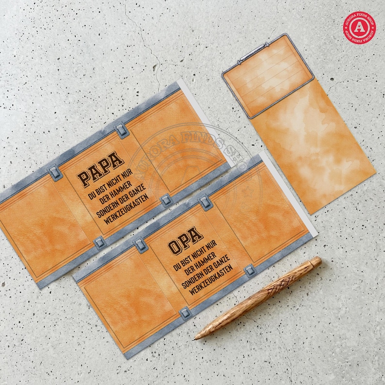May include: A set of orange and gray paper cards with text in German, including "PAPA" and "OPA". The cards have a toolbox design. A wooden pen lies on the surface. The cards are on a light gray surface.