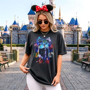 May include: Dark grey t-shirt with a vibrant graphic of Disney villains, including Maleficent and Cruella de Vil. The design features a crew neck and short sleeves. The shirt is a casual style.