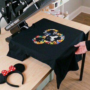 Peut inclure: T-shirt noir avec un motif color&eacute; en forme de t&ecirc;te de souris rempli de personnages de dessins anim&eacute;s. Un serre-t&ecirc;te &agrave; n&oelig;ud rouge et blanc &agrave; pois et une presse &agrave; chaud sont &eacute;galement visibles.