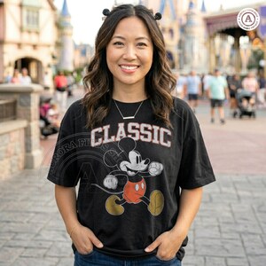 May include: Black t-shirt featuring a classic Mickey Mouse graphic. The word "CLASSIC" is printed across the top in white and red lettering. The shirt has short sleeves and a relaxed fit.