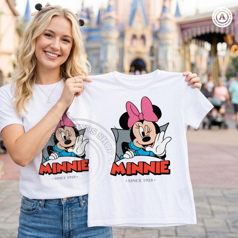 May include: White t-shirt featuring a Minnie Mouse graphic. The design includes Minnie's face with a pink bow, blue dress, and the word "MINNIE" in red, with "SINCE 1928" below. The shirt is held up by a person.