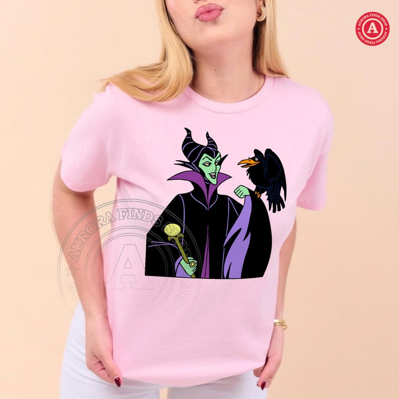May include: Pink t-shirt featuring a cartoon graphic of a character with green skin, black horns, and a black crow. The character is wearing a black and purple outfit and holding a golden staff. The shirt has a round neckline.