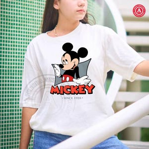 May include: White t-shirt featuring a Mickey Mouse graphic. The design includes Mickey in a black shirt, red shorts, and white gloves, with the word "MICKEY" in red and the text "SINCE 1928" below.