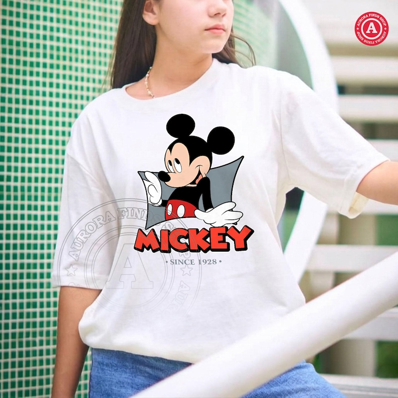 May include: White t-shirt featuring a Mickey Mouse graphic. The design includes Mickey in a black shirt, red shorts, and white gloves, with the word "MICKEY" in red and the text "SINCE 1928" below.
