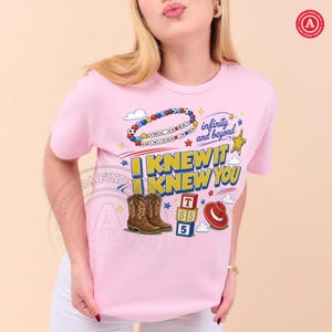 Puede incluir: Una camiseta rosa claro con un estampado gr&aacute;fico que incluye una pulsera de cuentas, un sombrero de vaquero, bloques de construcci&oacute;n y botas de vaquero marrones. El texto dice "YOU'VE GOT A FRIEND IN ME", "infinity and beyond" e "I KNEW IT I KNEW YOU".