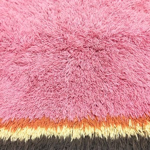 May include: A shaggy rug featuring a large section of pink fibers, followed by horizontal bands of orange, cream, and dark brown. The surface is composed of long, dense, and textured pile, creating a soft and voluminous appearance throughout the rug.