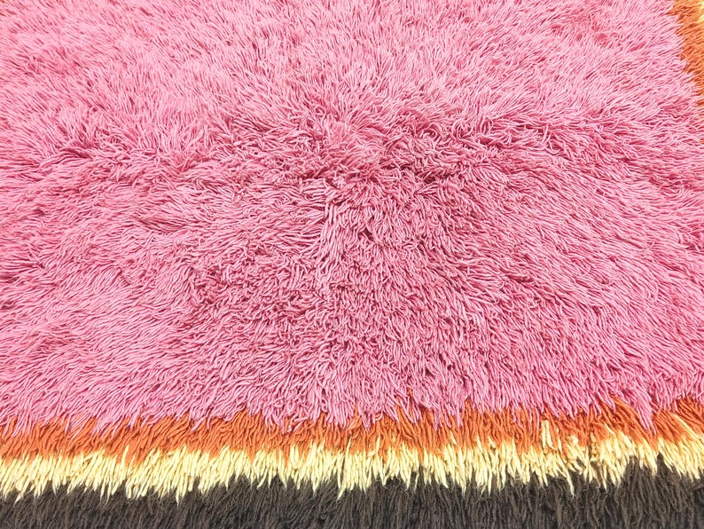 May include: A shaggy rug featuring a large section of pink fibers, followed by horizontal bands of orange, cream, and dark brown. The surface is composed of long, dense, and textured pile, creating a soft and voluminous appearance throughout the rug.