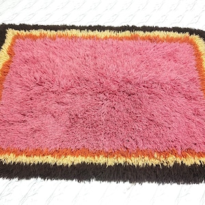 May include: A rectangular shag rug featuring a large central field of pink fibers surrounded by concentric borders of orange, yellow, and dark brown. The rug has a thick, high-pile texture throughout, with long, soft fibers creating a dense surface.
