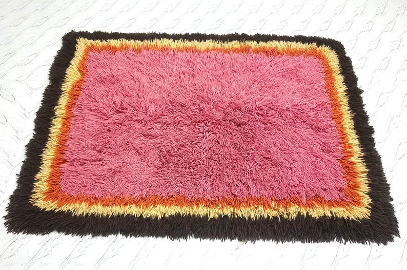 May include: A rectangular shag rug featuring a large central field of pink fibers surrounded by concentric borders of orange, yellow, and dark brown. The rug has a thick, high-pile texture throughout, with long, soft fibers creating a dense surface.