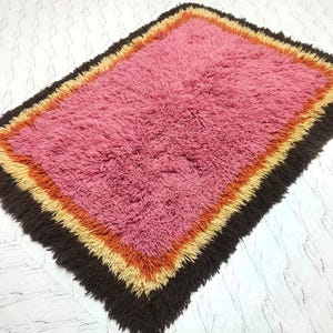 May include: A rectangular shag rug featuring a large central field of pink fibers surrounded by concentric borders of orange, yellow, and dark brown. The rug has a thick, textured pile with a shaggy appearance and a multi-colored layered design.