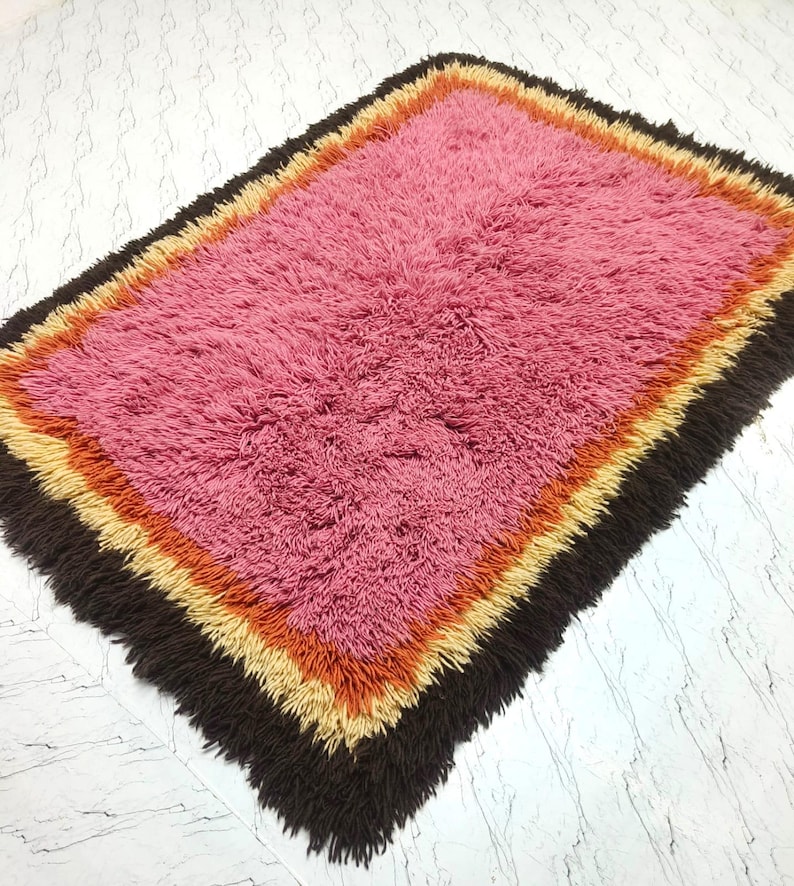 May include: A rectangular shag rug featuring a large central field of pink fibers surrounded by concentric borders of orange, yellow, and dark brown. The rug has a thick, textured pile with a shaggy appearance and a multi-colored layered design.
