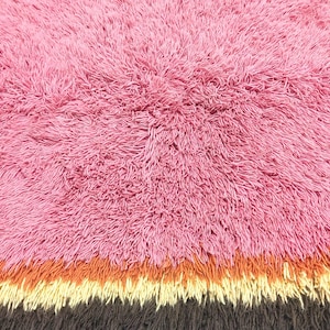 May include: A shaggy rug featuring a large section of pink fibers, followed by horizontal bands of orange, cream, and dark brown. The texture is dense and long-piled, creating a soft, uneven surface across the entire rectangular area of the rug.