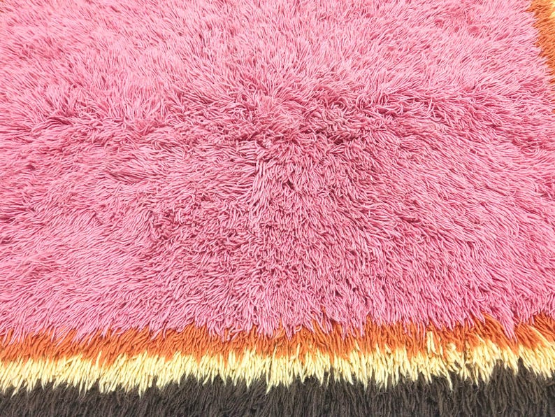 May include: A shaggy rug featuring a large section of pink fibers, followed by horizontal bands of orange, cream, and dark brown. The texture is dense and long-piled, creating a soft, uneven surface across the entire rectangular area of the rug.