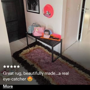 May include: A rectangular, shaggy area rug featuring a gradient of dark brown, purple, and cream fibers. The rug is positioned on a light-colored floor beneath a black slatted bench holding a pink handbag, a light blue handbag, and a baseball cap.