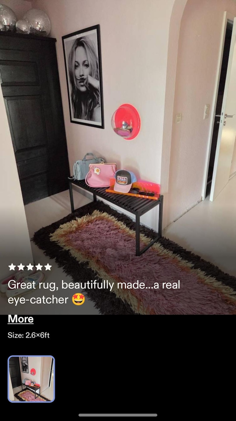 May include: A rectangular, shaggy area rug featuring a gradient of dark brown, purple, and cream fibers. The rug is positioned on a light-colored floor beneath a black slatted bench holding a pink handbag, a light blue handbag, and a baseball cap.