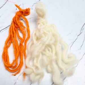 Pode incluir: Dois feixes de fio, um laranja brilhante e outro branco sujo, cada um com uma se&ccedil;&atilde;o superior com n&oacute;s e longos fios soltos e ondulados pendurados. O fio laranja parece mais fino e mais torcido, enquanto o fio branco &eacute; mais espesso.