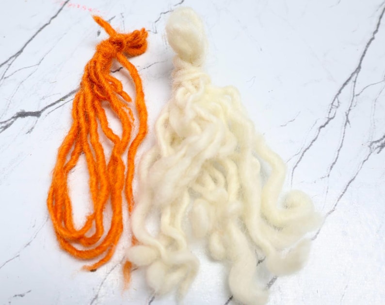 Pode incluir: Dois feixes de fio, um laranja brilhante e outro branco sujo, cada um com uma se&ccedil;&atilde;o superior com n&oacute;s e longos fios soltos e ondulados pendurados. O fio laranja parece mais fino e mais torcido, enquanto o fio branco &eacute; mais espesso.