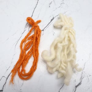 Pode incluir: Dois feixes de fio, um em uma cor laranja vibrante e o outro em uma cor creme esbranqui&ccedil;ada. O fio laranja consiste em v&aacute;rios fios longos e finos amarrados no topo, enquanto o fio creme &eacute; mais grosso, texturizado e reunido de forma solta.