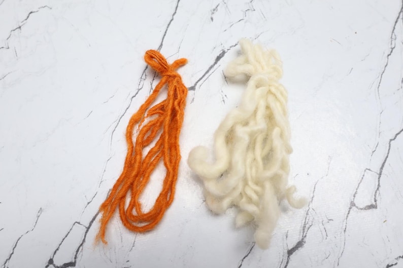 Pode incluir: Dois feixes de fio, um em uma cor laranja vibrante e o outro em uma cor creme esbranqui&ccedil;ada. O fio laranja consiste em v&aacute;rios fios longos e finos amarrados no topo, enquanto o fio creme &eacute; mais grosso, texturizado e reunido de forma solta.
