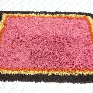 May include: A rectangular shag rug featuring a large central field of pink fibers surrounded by concentric borders of orange, yellow, and dark brown. The rug has a thick, high-pile texture throughout, creating a dense and soft appearance.