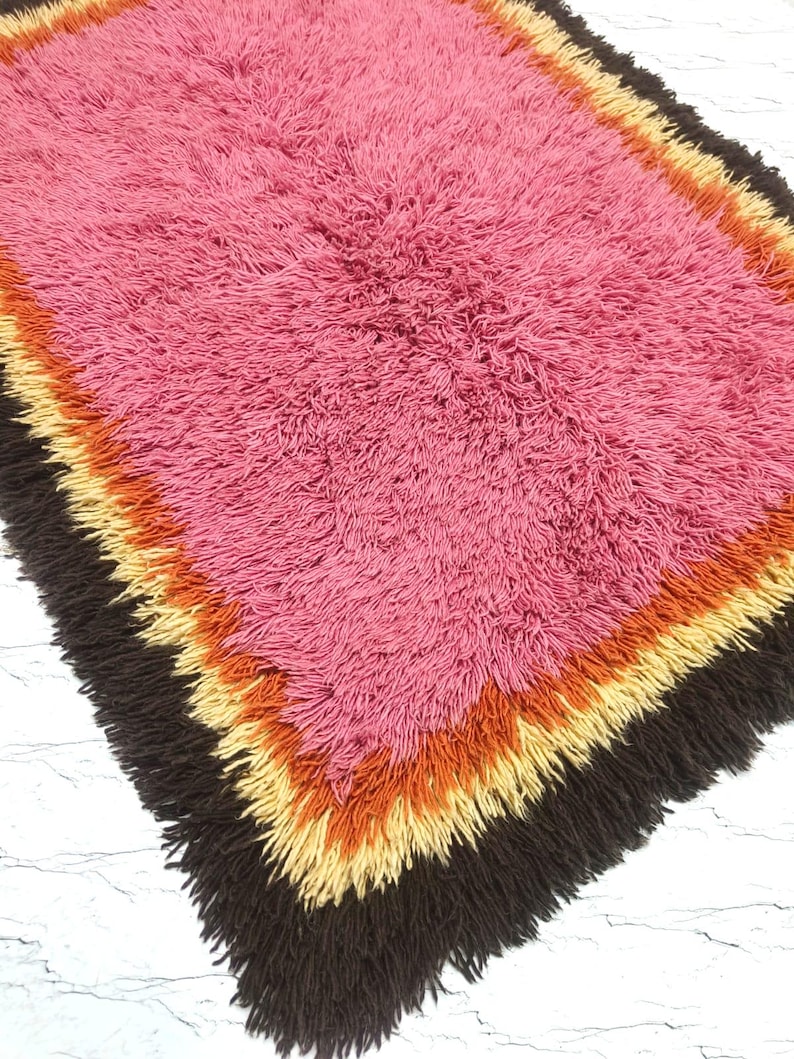 May include: A rectangular shag rug featuring a large central field of vibrant pink surrounded by concentric borders of orange, yellow, and dark brown. The rug has a thick, high-pile texture with long, soft fibers that create a dense, plush appearance.