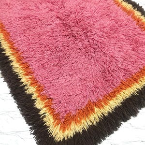 May include: A rectangular shag rug featuring a large central field of pink, surrounded by concentric borders of orange, yellow, and dark brown. The rug has a thick, high-pile texture with long, shaggy fibers throughout the entire surface area.