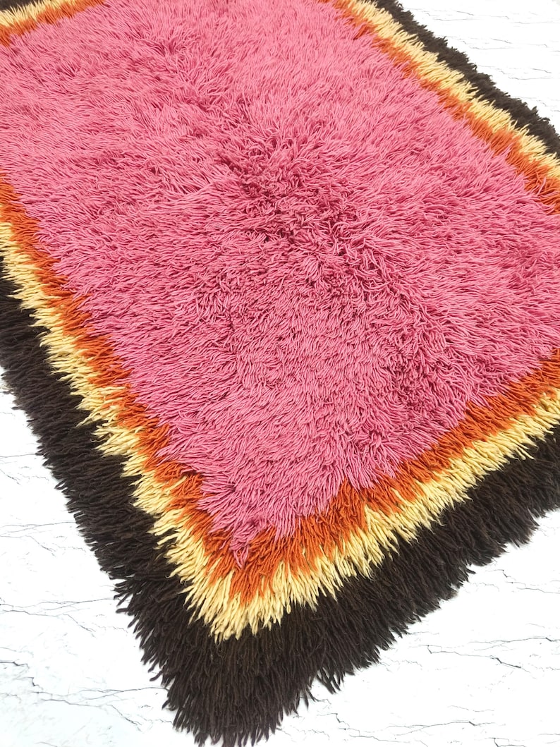 May include: A rectangular shag rug featuring a large central field of pink, surrounded by concentric borders of orange, yellow, and dark brown. The rug has a thick, high-pile texture with long, shaggy fibers throughout the entire surface area.