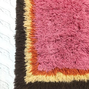 May include: A rectangular shag rug featuring a large central field of vibrant pink, surrounded by concentric borders in shades of bright orange, pale yellow, and dark brown. The rug has a thick, high-pile texture with long, soft fibers throughout.