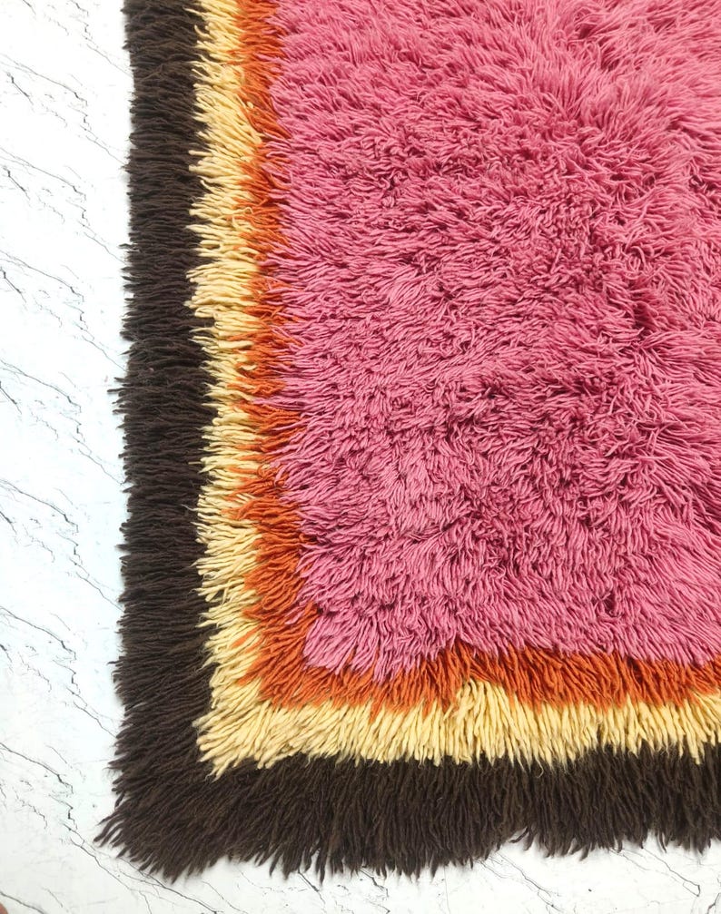 May include: A rectangular shag rug featuring a large central field of vibrant pink, surrounded by concentric borders in shades of bright orange, pale yellow, and dark brown. The rug has a thick, high-pile texture with long, soft fibers throughout.