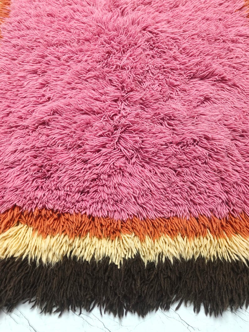 May include: A high-pile shag rug featuring a large section of vibrant pink fibers at the top, followed by horizontal bands of burnt orange, cream, and dark brown at the bottom. The texture is dense, shaggy, and uniform across the entire surface.