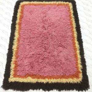 May include: A rectangular shag rug featuring a large central pink section surrounded by concentric borders in orange, light yellow, and a dark brown outer edge. The rug has a thick, textured pile throughout, creating a soft and dense appearance.