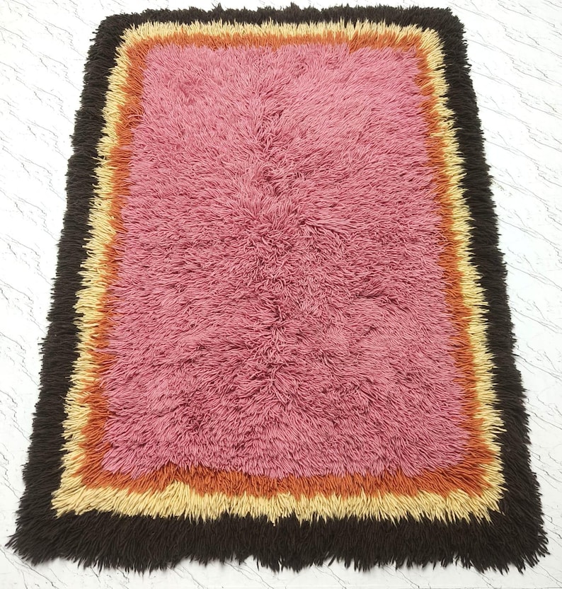 May include: A rectangular shag rug featuring a large central pink section surrounded by concentric borders in orange, light yellow, and a dark brown outer edge. The rug has a thick, textured pile throughout, creating a soft and dense appearance.