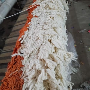 Pode incluir: Uma pe&ccedil;a t&ecirc;xtil longa e retangular com uma textura de pelo grosso e felpudo. A superf&iacute;cie &eacute; dividida em duas sec&ccedil;&otilde;es de cores distintas, com um lado composto por la&ccedil;os de fio laranja brilhante e o outro por fio de cor creme esbranqui&ccedil;ado.