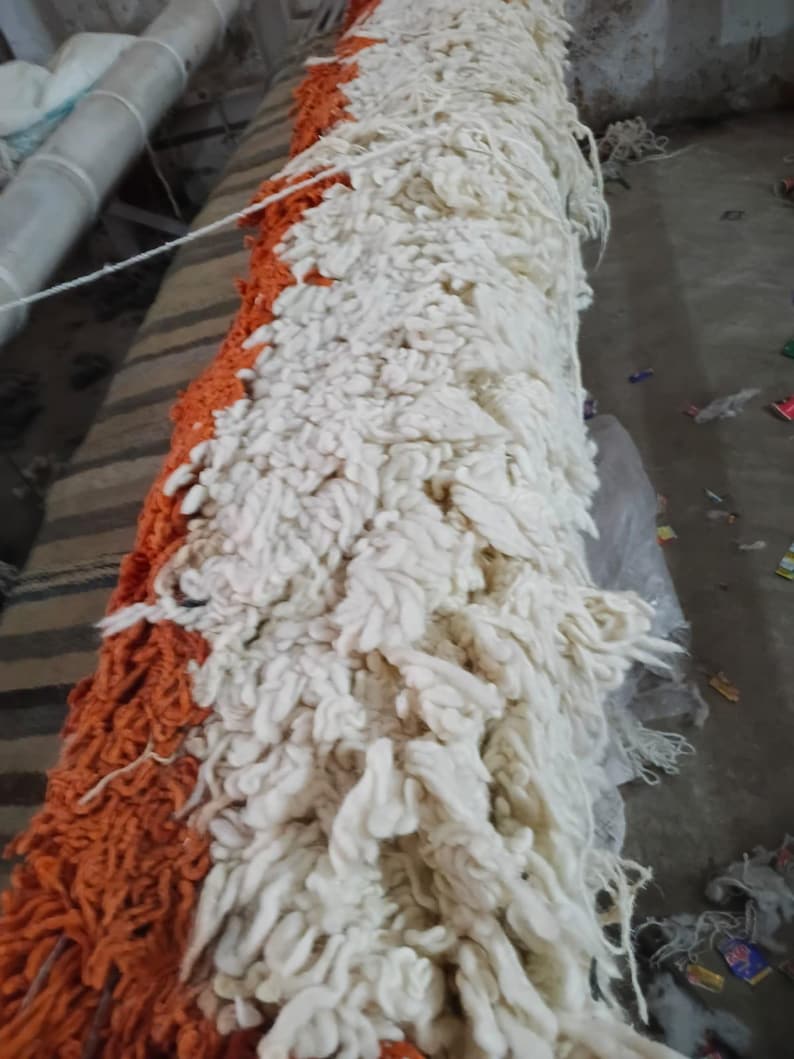 Pode incluir: Uma pe&ccedil;a t&ecirc;xtil longa e retangular com uma textura de pelo grosso e felpudo. A superf&iacute;cie &eacute; dividida em duas sec&ccedil;&otilde;es de cores distintas, com um lado composto por la&ccedil;os de fio laranja brilhante e o outro por fio de cor creme esbranqui&ccedil;ado.